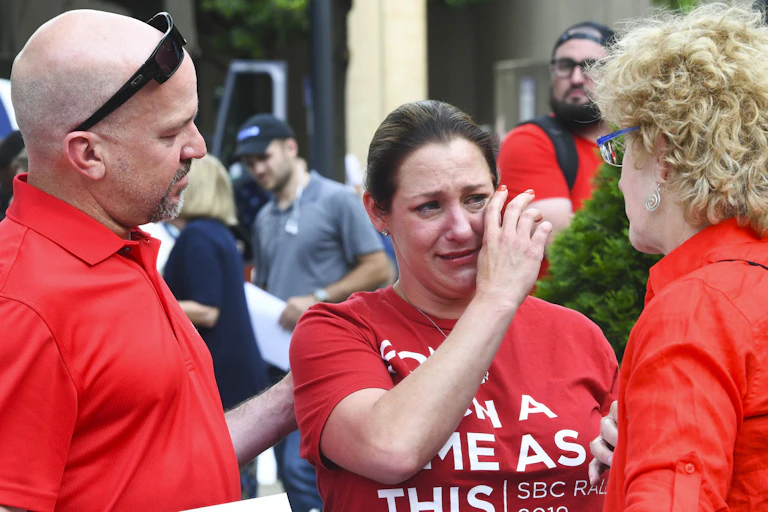 A woman dressed in a red T-shirt wipes her tears as her two friends standing next to her comfort her.