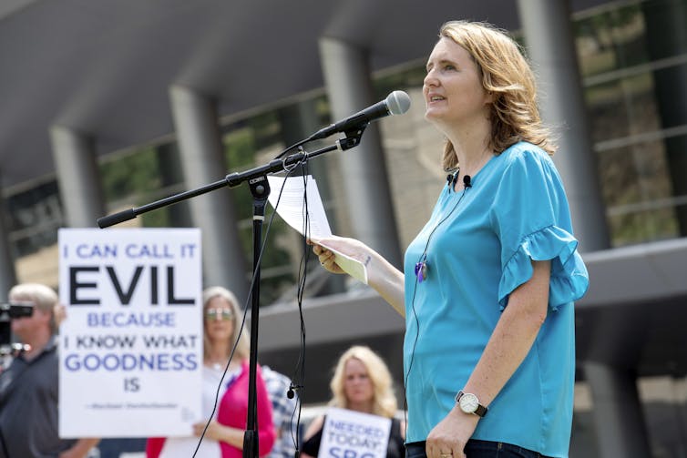 A woman wearing a blue shirt speaking at a microphone, with a poster by her side that says 'I can call it evil because I know what goodness is.'