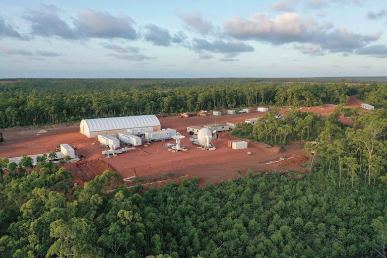 An aerial photograph of a hangar and other buildings on red earth in a clearing in the middle of a forest.