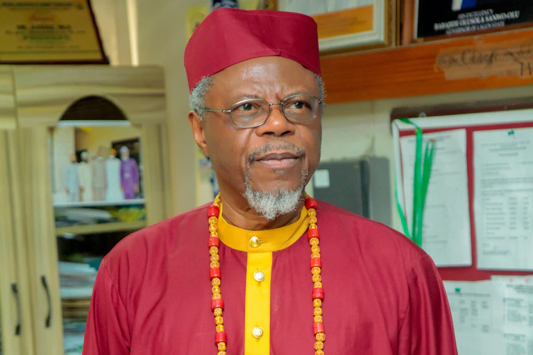 A man with greying beard wears red traditional attire and glasses, looking into the distance.