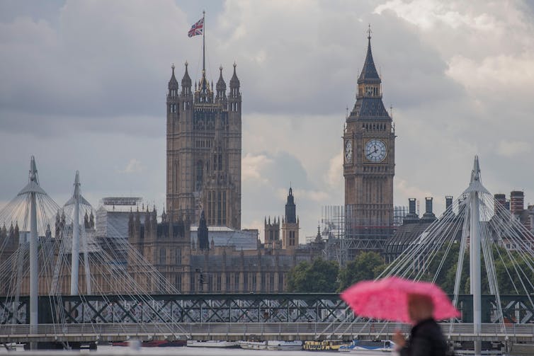 A person carrying a pink umbrella walks in the foreground, while the houses of Parliament are visible in the background against a grey sky