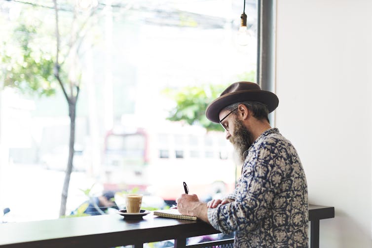 A man with a beard dressed stylishly in a brimmed hat and glasses sits at a window-facing counter in a coffee shop and writes by hand in a journal