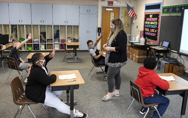 A teacher speaks with students who are raising their hands.