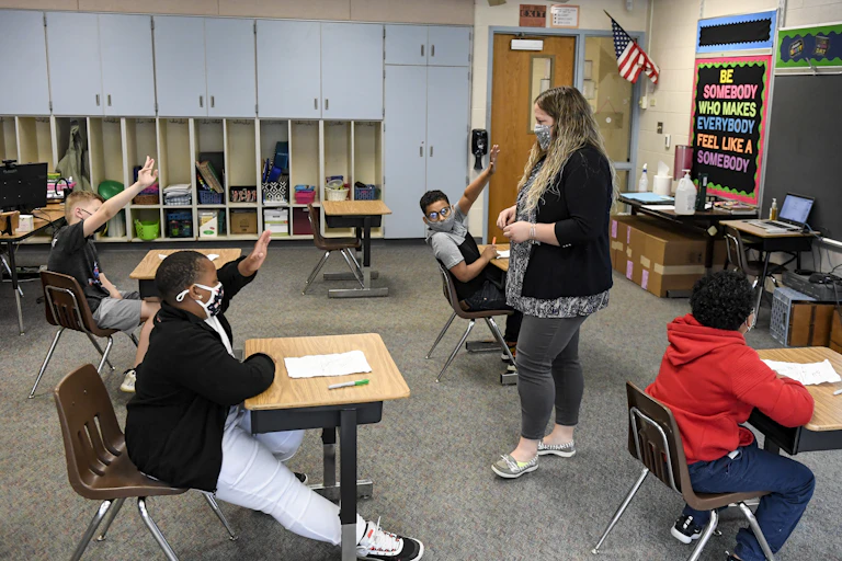 A teacher speaks with students who are raising their hands.