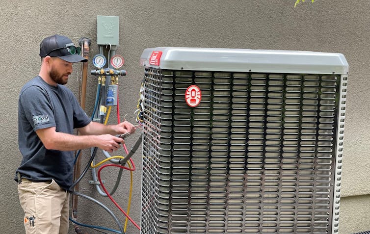 A worker in ballcap and short sleeves installs a large hat pump, hooking up hoses next to a house.