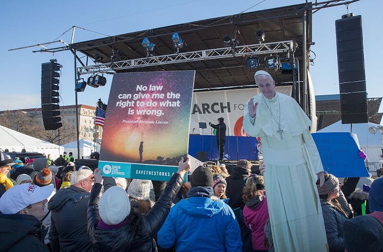 People at a rally hold a life-size illustration of Pope Francis in front of an outdoor stage.