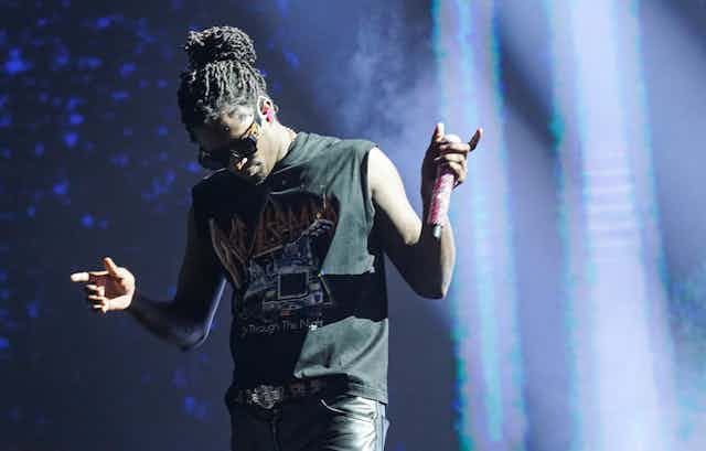 A Black man with dreadlocks wearing a black T-shirt holds a microphone while performing on stage.