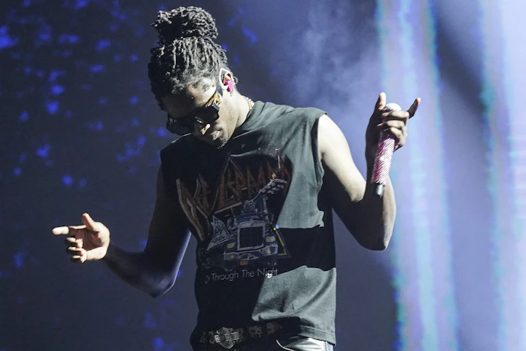 A Black man with dreadlocks wearing a black T-shirt holds a microphone while performing on stage.