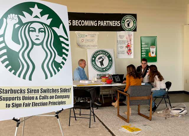 A poster depicting a depiction of the Starbucks mermaid logo with a raised fist stands near baristas sitting and talking about labor organizing 