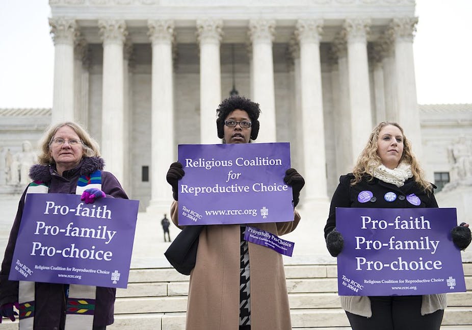 Three women in winter coats hold purple signs on the steps of the Supreme Court. 
