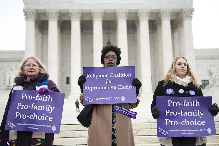 Three women in winter coats hold purple signs on the steps of the Supreme Court.