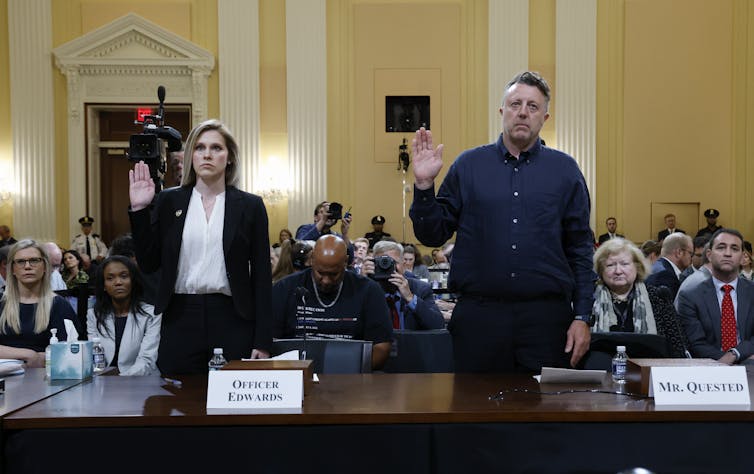 A solemn woman and man stand and raise their right hand as they are sworn in to testify.