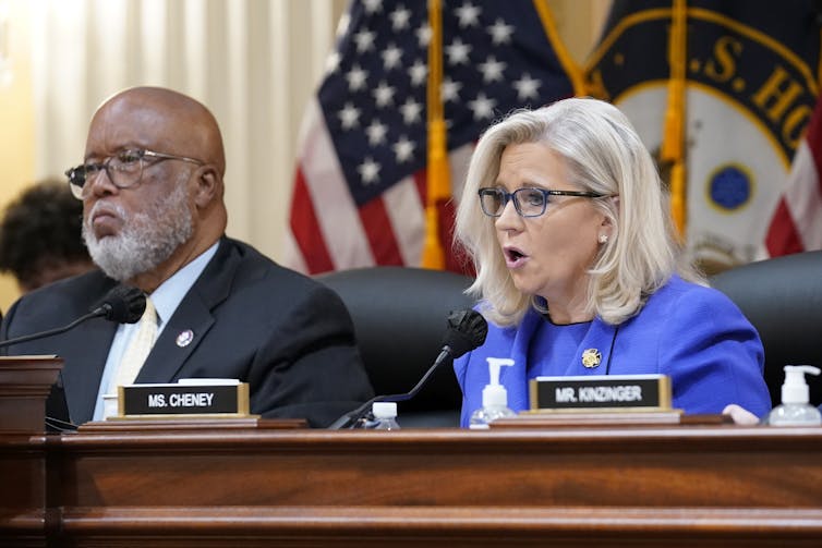 A woman in a blue jacket and wearing glasses is talking at a large desk in front of the American flag; next to her is a man in a suit and tie.