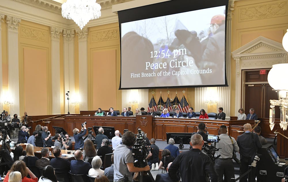 A video is projected on a screen above a committee of people, reading 12:54 pm Peace Circle, First Breach of the Capitol Grounds.