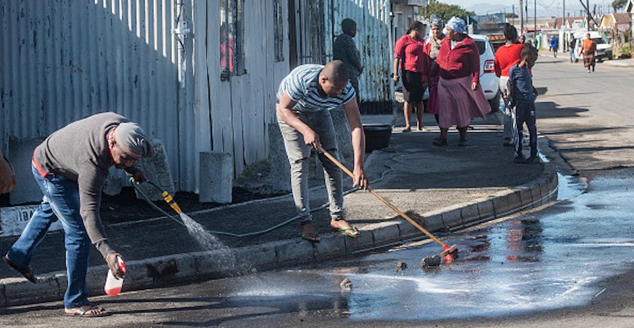 A man hoses down a street while another sweeps with a broom to remove blood from a tarred rowd, while few women watch.
