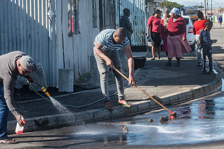 A man hoses down a street while another sweeps with a broom to remove blood from a tarred rowd, while few women watch.