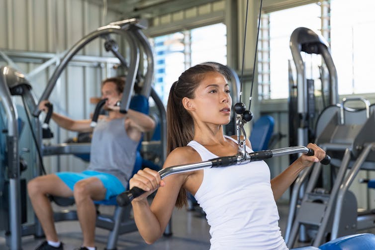 A woman in the gym performs a lat pulldown. A man behind her is using a machine to perform a chest press.