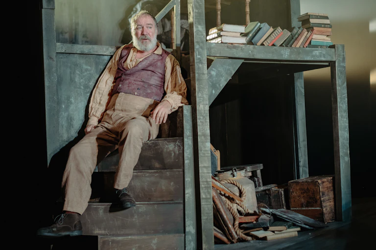 A scene from the play Translations showing a man in late 19th century dress sitting on some wooden stairs by a pile of books.