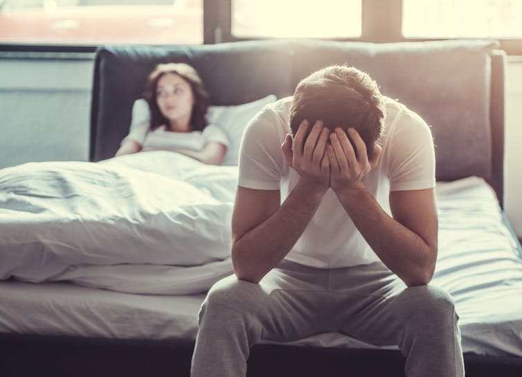 A man sits at the end of the bed with his head in his hands, the woman is leaning back in bed looking upset.