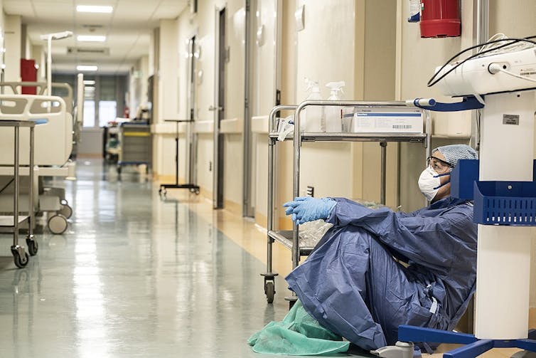 A nurse in protective gear sits on the floor in an empty hallway.
