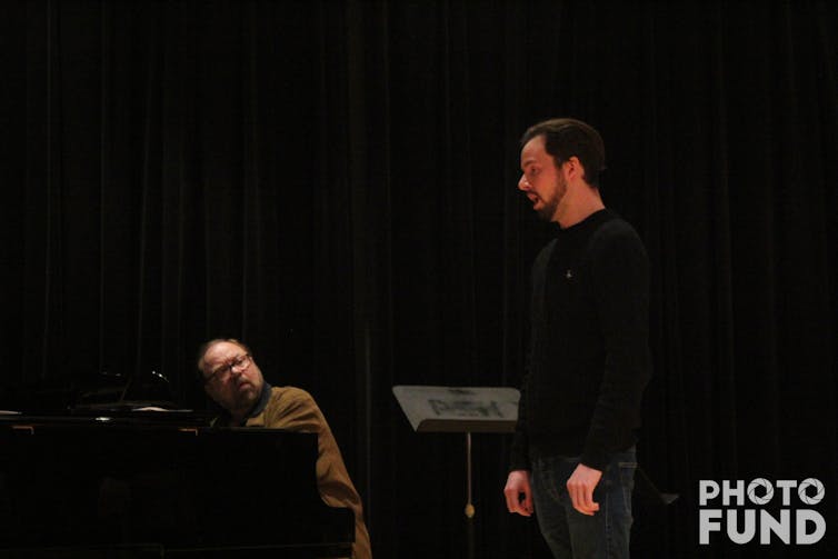 A man standing up with a music stand behind him, and another man sitting down and looking at him