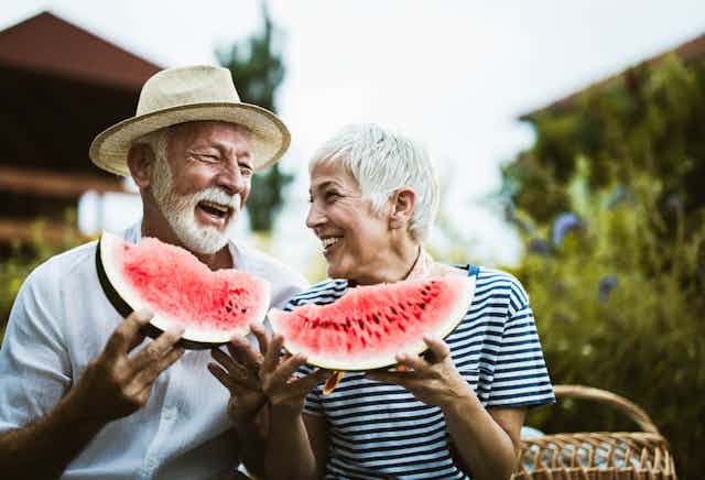 An elderly man wearing a brimmed hat and an elderly woman smile as they eat watermelon