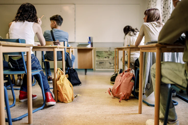 High school students are seen from the back as they sit at desks looking to the left.