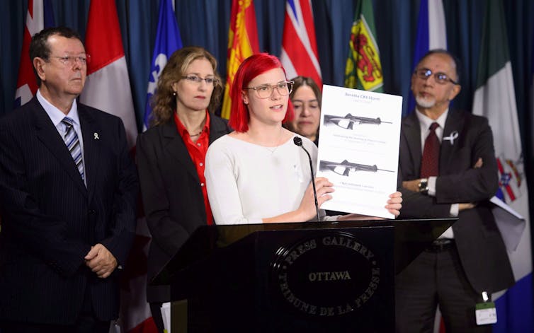 A red-haired woman holds up a placard with assault-style weapons on it.