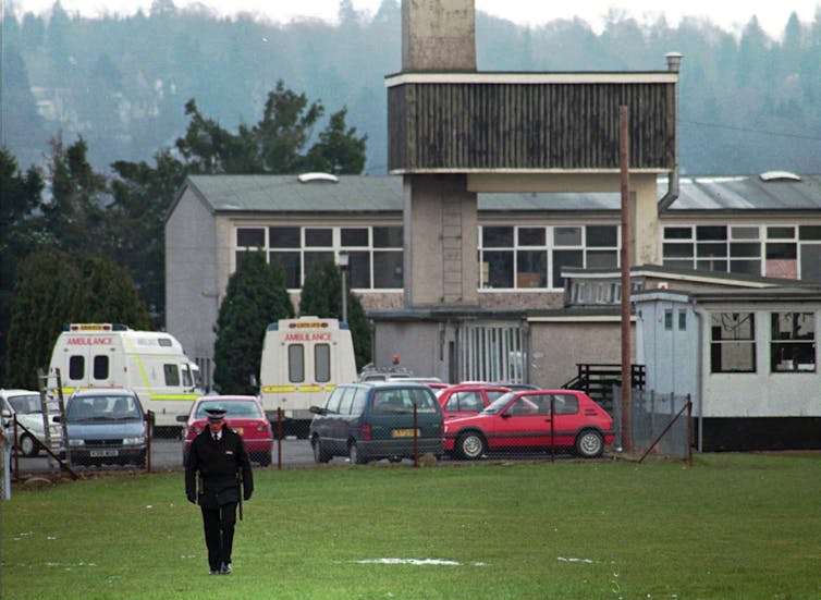 A police officer walks on the grass outside a school. Ambulances sit in the parking lot behind him.