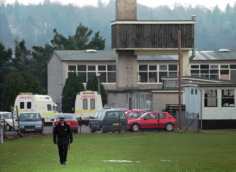 A police officer walks on the grass outside a school. Ambulances sit in the parking lot behind him.