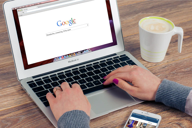 Hands resting on a laptop keyboard with a search for 'headache, sneezing, itchy eyes' on the screen, with a mug on the table