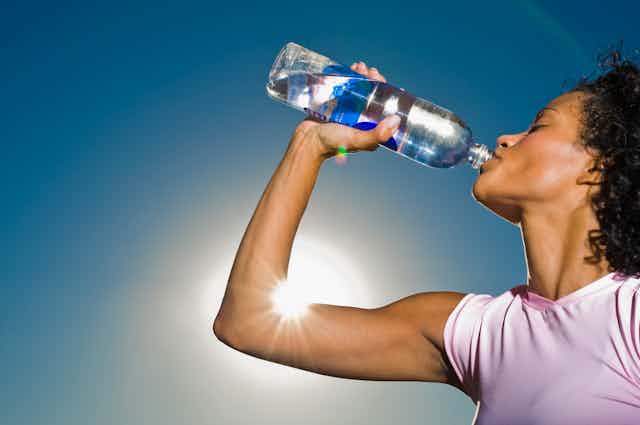 A woman, standing outside in the sunshine, drinks water from a plastic bottle.