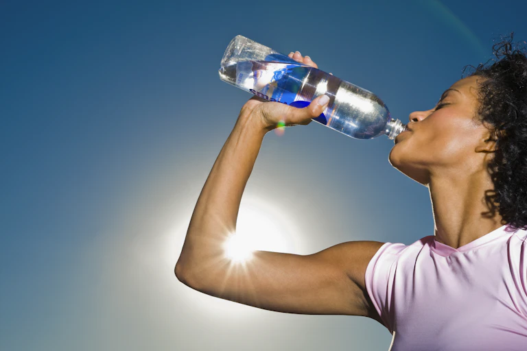 A woman, standing outside in the sunshine, drinks water from a plastic bottle.