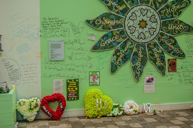 A green-painted section of wall with a floral mosaic, floral tributes and written messages.