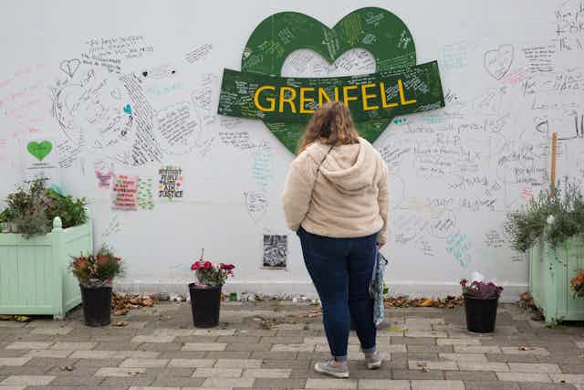 A woman stands with her back to the camera, looking at a green heart and other messages on a wall.