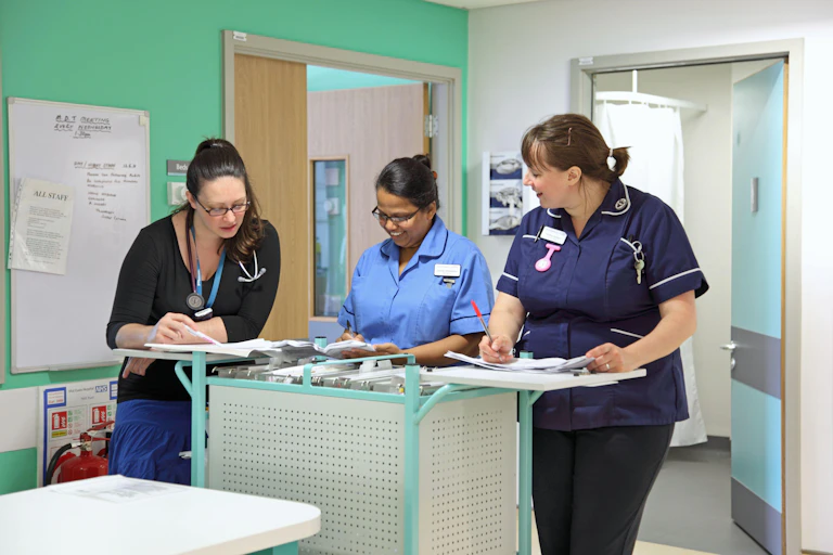 Three nurses stand at a counter in a green hospital room.