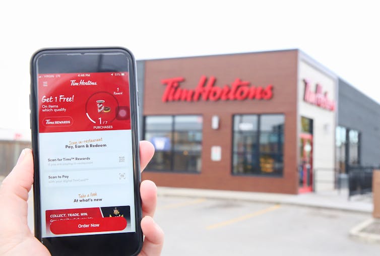 A hand holds a mobile screen showing the Tim Hortons app. There's a Tim Hortons restaurant in the background
