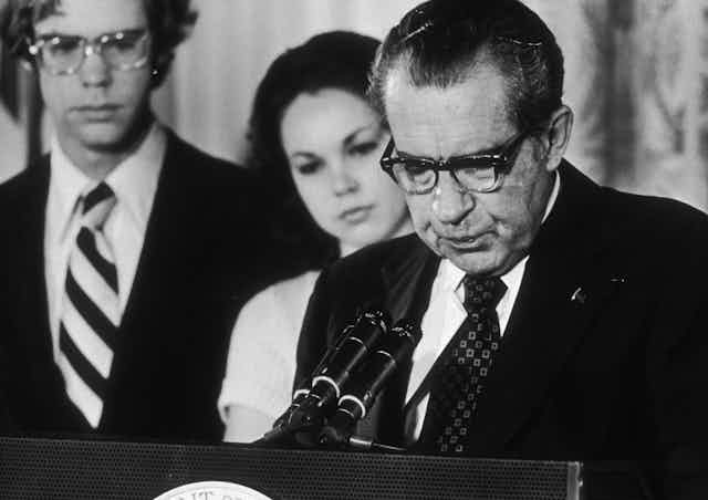 A dark haired man in glasses, wearing a dark suit jacket, standing at some microphones and talking. He's standing next to a young woman and man.