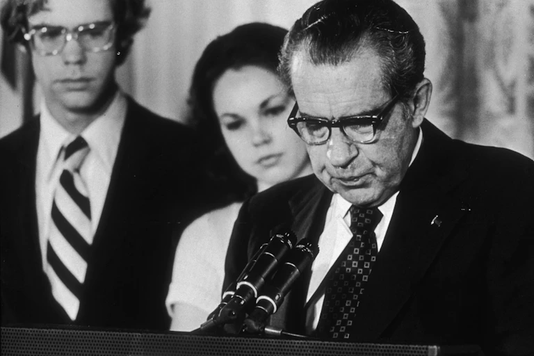 A dark haired man in glasses, wearing a dark suit jacket, standing at some microphones and talking. He's standing next to a young woman and man.