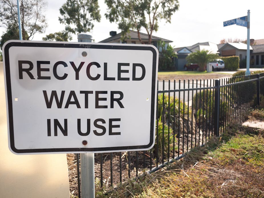A sign reads "Recycled water in use" in black letters against a white background, alongside some grass and shrubs