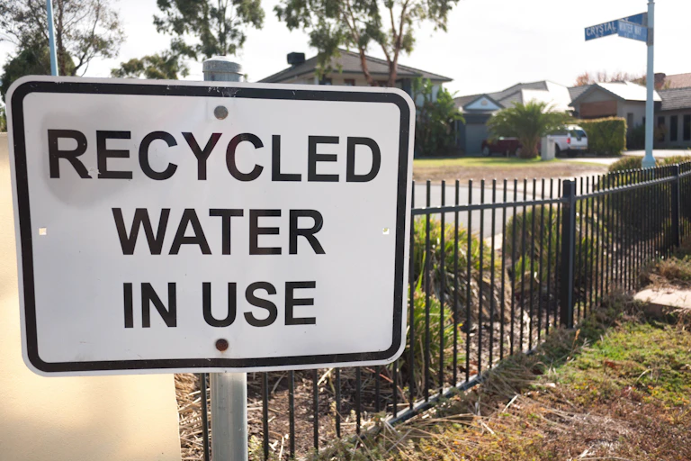A sign reads "Recycled water in use" in black letters against a white background, alongside some grass and shrubs