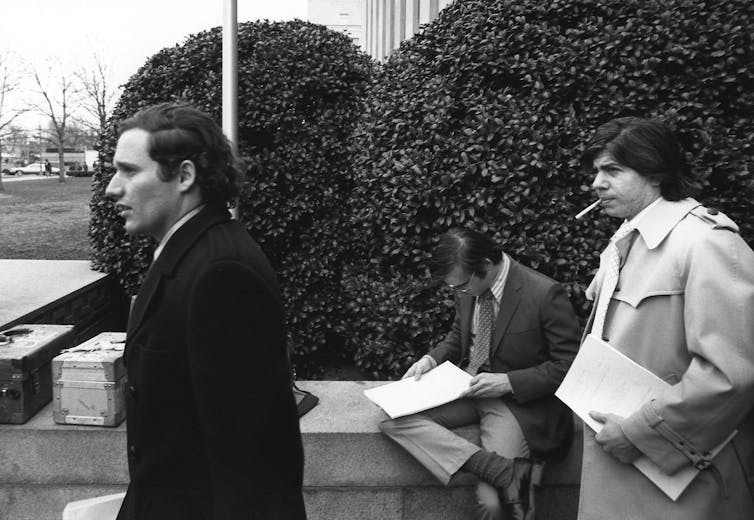 Two men, one in a topcoat and one in a raincoat, walk away from a building. One is carrying a file folder.