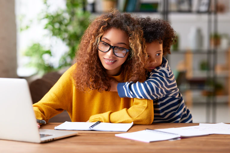 A mother works on her laptop while a child hugs her from behind