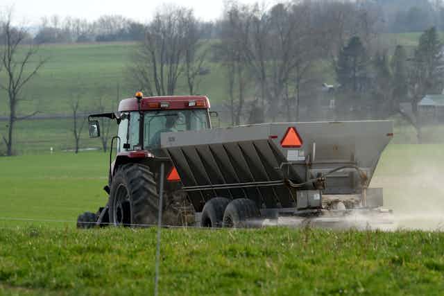 A tractor drives across a field with fertilizer pouring out of an attached hopper.
