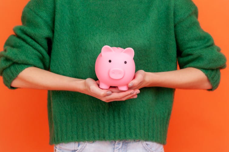 Torso of a woman in a green sweater holding a piggy bank in front of her, against an orange wall