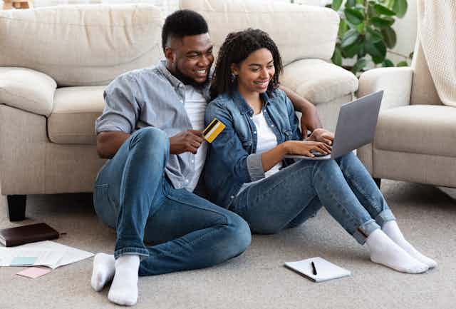 A young couple sitting on the floor, looking happily at a laptop and holding a credit card