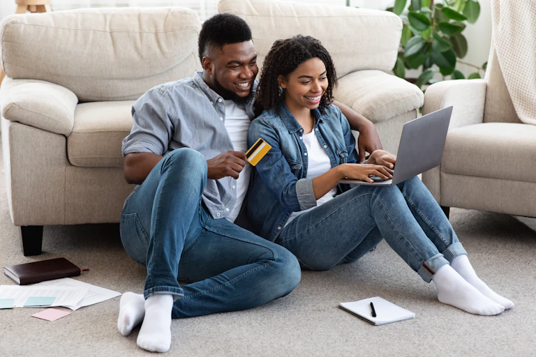 A young couple sitting on the floor, looking happily at a laptop and holding a credit card