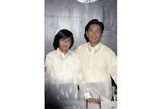 Two men in cream-coloured clothing, one a teenaged boy, smile at the camera.