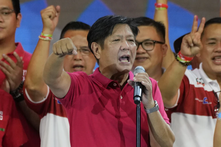 A man in a red polo shirt speaks into a microphone with his fist raised.