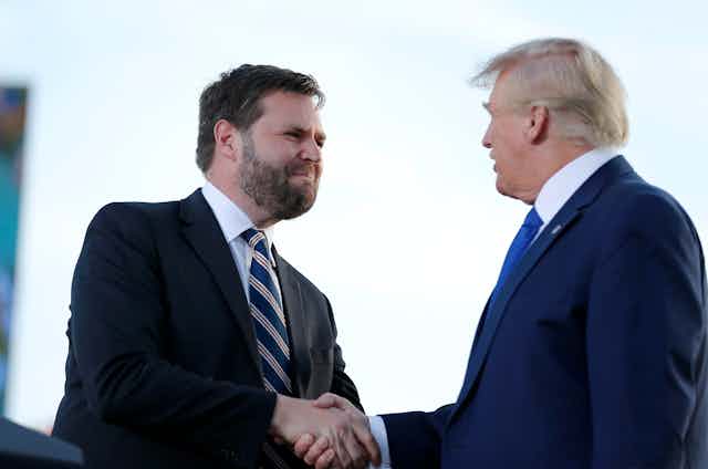 A younger man with dark hair and a beard shakes hands of an older man with light hair. Both are wearing dark blazers and ties.